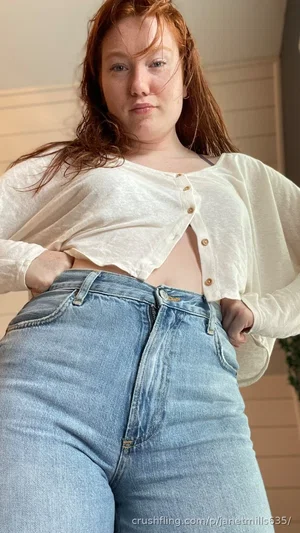 Woman with red hair in white cropped cardigan and high-waisted jeans, standing indoors.
