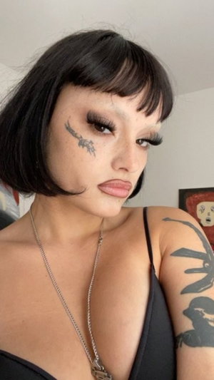 Woman with black bob, tattoos, and black top in indoor setting.