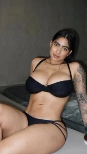 Woman in black lingerie posing indoors, tattoo on arm, dark hair.