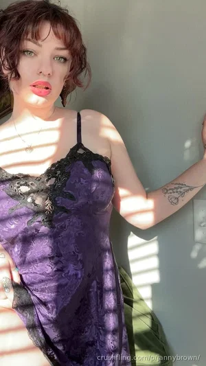 Woman in purple lace dress with tattoos, posing indoors near a wall.