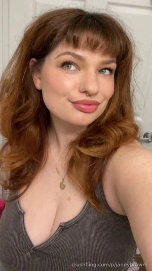Woman with wavy brown hair and bangs wearing a gray tank top, smiling in a bathroom selfie.