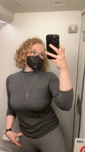 Woman with curly blonde hair in gray long-sleeve top and leggings, taking mirror selfie in small