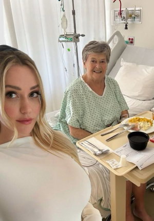 Blonde woman in white top takes selfie with older woman in hospital gown at bedside.