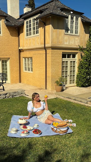 Woman in white top and skirt having picnic on lawn in front of stone house.
