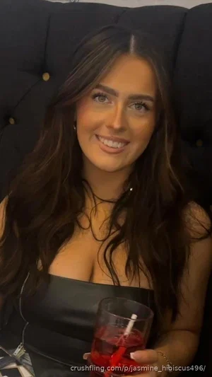 Woman with long dark hair in black strapless top holding drink, seated on dark tufted couch.