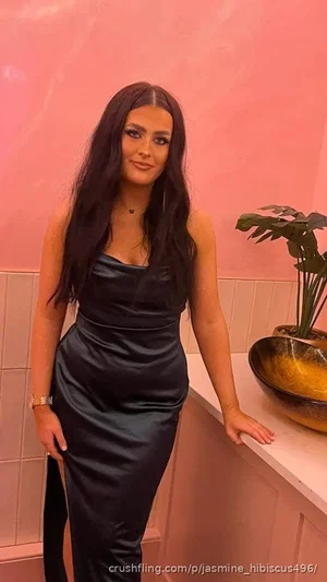 Woman in black satin dress with long dark hair, standing in pink-tiled room with plant.