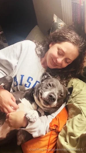 Woman with curly hair in a white sweatshirt holding a gray and white dog on a bed.