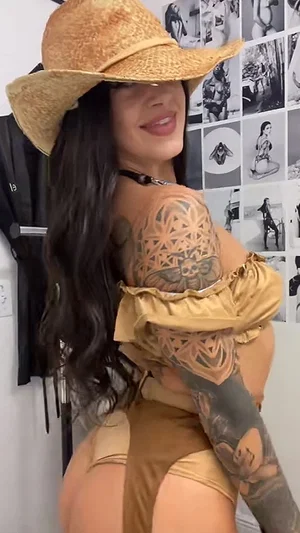 Woman with long dark hair and tattoos wears gold outfit and straw hat, posing indoors.