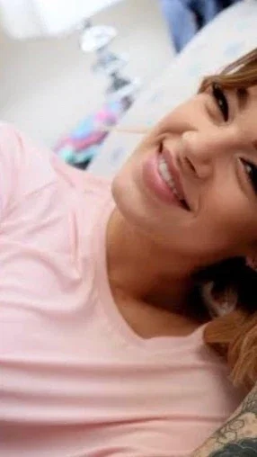 Woman with tattoo on arm wearing pink shirt, smiling in indoor setting.