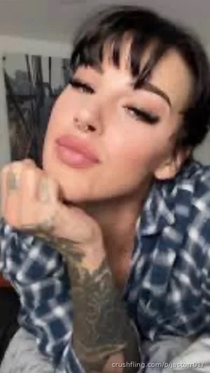 Woman with dark hair and tattoos wears a plaid shirt, resting chin on hand indoors.