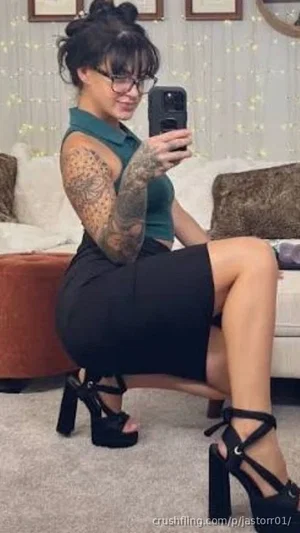 Woman with tattoos in green top and black skirt, sitting on couch, taking mirror selfie.
