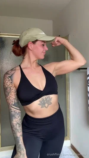 Woman in black sports bra and leggings flexing arm, tattoos visible, wearing cap, indoor setting.