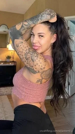 Woman with long dark hair and full sleeve tattoos wears pink crop top and black pants indoors.