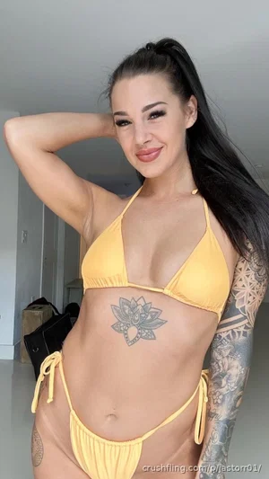 Woman in yellow bikini with tattoos, posing indoors, long dark hair, smiling.