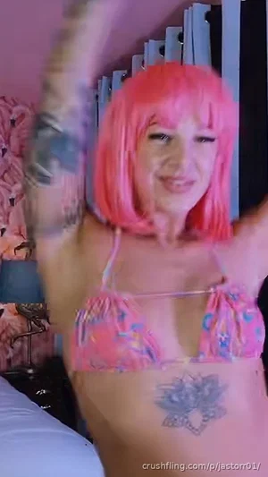 Woman with pink wig and tattoos wears floral bikini top in bedroom.