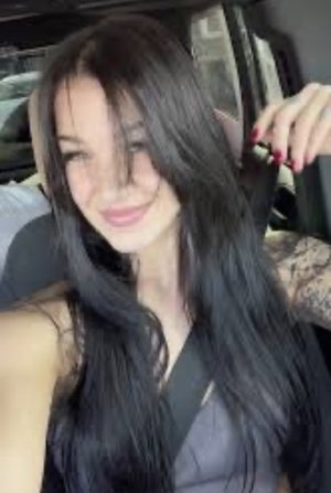 Woman with long dark hair and tattoos in a car, wearing a dark top.