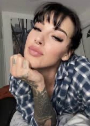 Woman with tattoos and dark hair in plaid shirt, resting chin on hand indoors.
