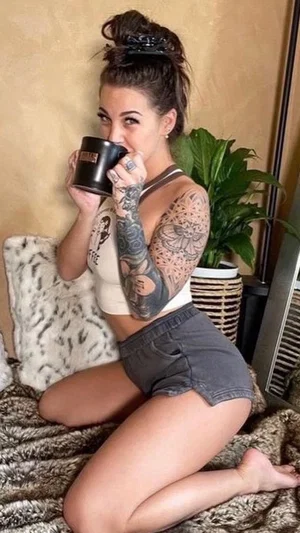 Woman with tattoos in beige top and shorts, kneeling on bed holding mug.