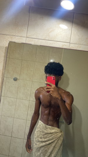 Shirtless man with towel wrapped around waist, taking mirror selfie in tiled bathroom.