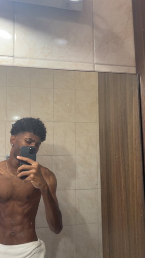 Shirtless man with curly hair taking mirror selfie in tiled bathroom, wearing white towel around