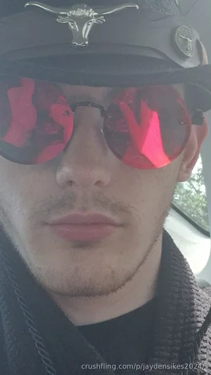 Man with beard wearing black hat and red sunglasses, indoors in a car.