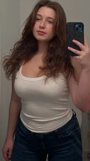 Woman with long brown hair in white tank top and jeans taking mirror selfie indoors.