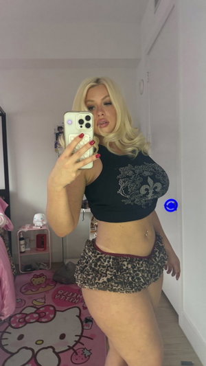 Blonde woman in crop top and shorts taking mirror selfie in bedroom.