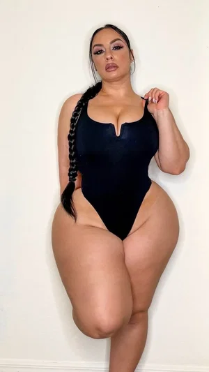 Woman in black bodysuit posing against white wall, long braided hair, curvy figure.