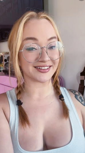 Blonde woman with glasses and braids in a light blue tank top, smiling indoors.