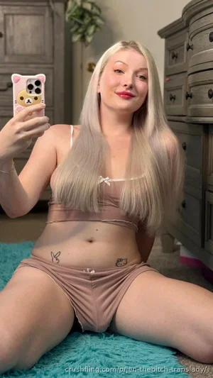 Woman with long blonde hair in pink lingerie sitting on blue rug, holding phone for mirror selfie.