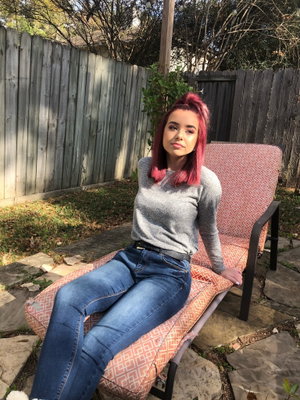 Woman with pink hair in gray sweater and jeans sitting on outdoor lounge chair.