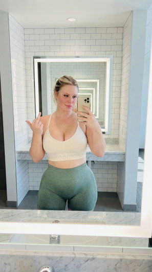 Woman in white sports bra and green leggings taking mirror selfie in tiled bathroom.