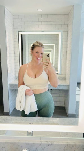 Woman in beige sports bra and green leggings takes mirror selfie in bathroom holding towel.