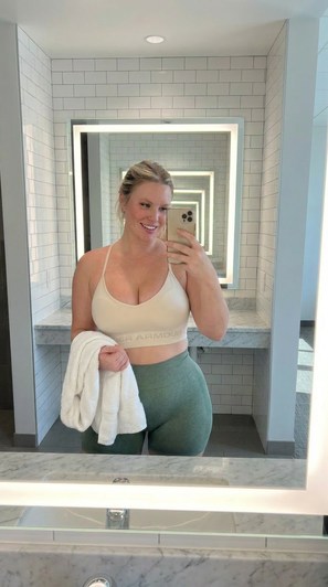 Woman in beige sports bra and green leggings holding towel, taking mirror selfie in tiled bathroom.