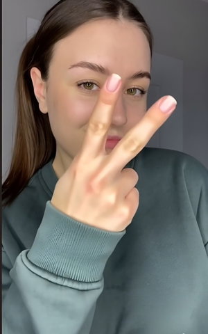 Woman with ponytail in green hoodie makes peace sign indoors.