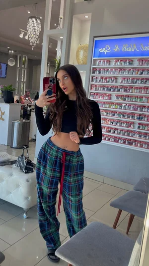Woman with long dark hair in black crop top and plaid pants taking mirror selfie in salon.