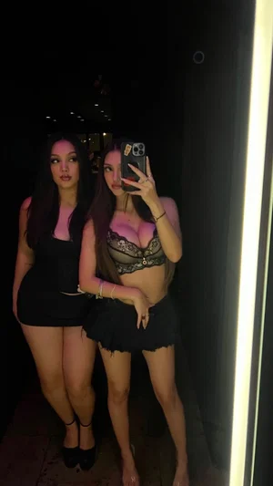 Two women in lingerie and short skirts pose together in a dimly lit room, one holding a phone for a