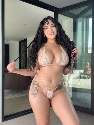 Woman with curly hair in beige bikini posing indoors near glass doors.
