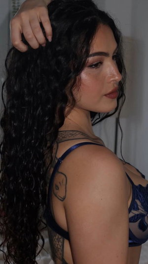 Woman with long dark curly hair, wearing blue lace lingerie, showing tattoos on back and shoulder,