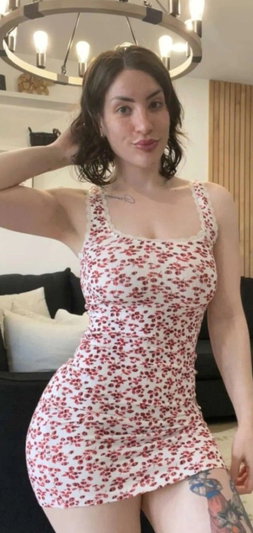 Woman in floral dress posing indoors, tattoo on thigh, dark hair, smiling.