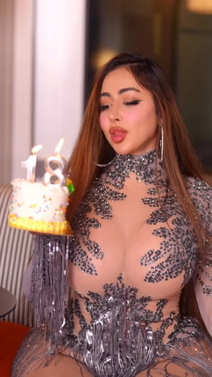 Curvy brunette in sheer sequined dress holds cake indoors.
