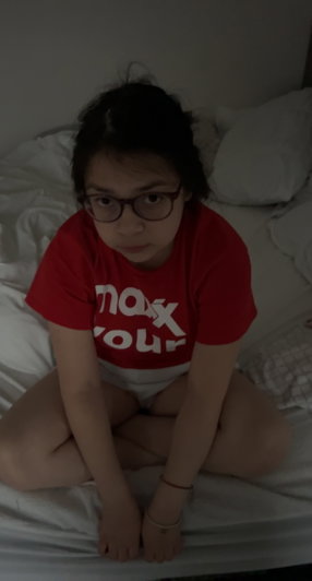 Asian woman with glasses in red shirt sitting on bed.