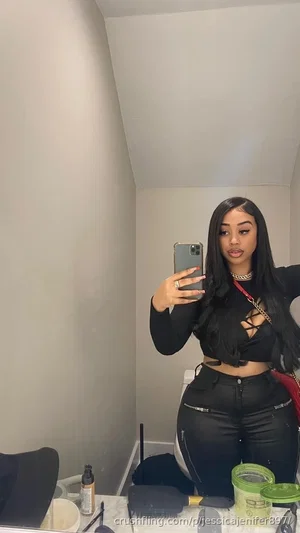 Woman with long black hair in black crop top and leather pants, taking mirror selfie in bathroom.