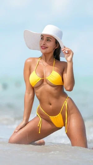 Woman in yellow bikini and white hat posing on beach, kneeling in shallow water.