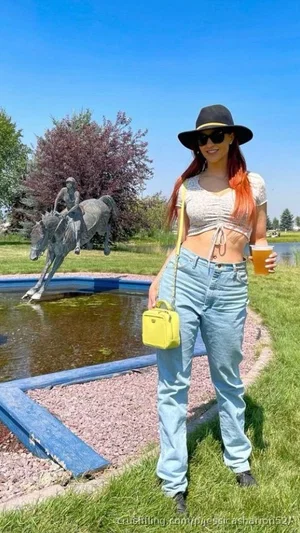 Woman with red hair in a crop top and jeans, holding a drink, standing outdoors near a statue.