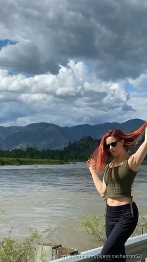 Woman with red hair in green crop top and black pants, posing by a river with mountains in