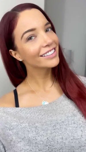 Woman with red hair wearing gray sweater and black strap, smiling indoors.