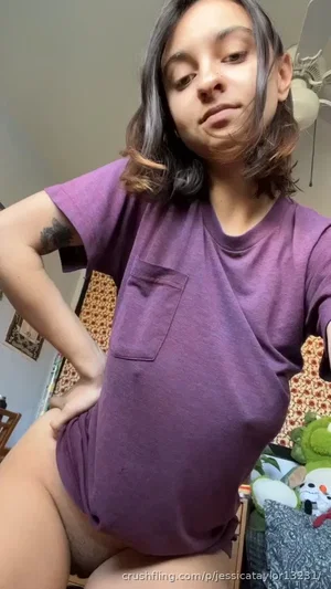 Woman with short dark hair in purple t-shirt, sitting indoors, hand on hip.