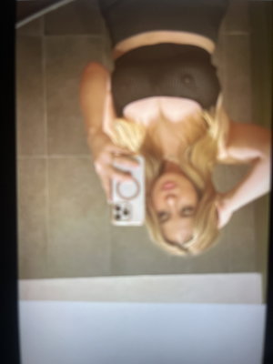 Blonde woman in black lingerie taking mirror selfie in tiled room.