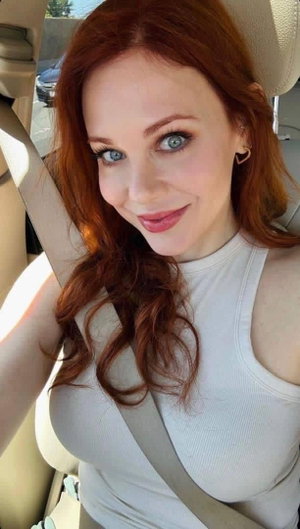 Woman with red hair and blue eyes wearing white tank top in car.